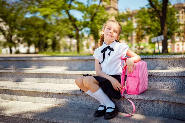 Merdivenlerde oturan ilkokul öğrencisi elini çantanın üzerine koydu ve gülümseyerek kameraya baktı. Açık havada binanın yakınında sırt çantası olan kız. Derslerin başlangıcı. Sonbaharın ilk günü