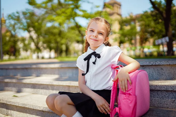 Merdivenlerde oturan ilkokul öğrencisi elini çantanın üzerine koydu ve gülümseyerek kameraya baktı. Açık havada binanın yakınında sırt çantası olan kız. Derslerin başlangıcı. Sonbaharın ilk günü