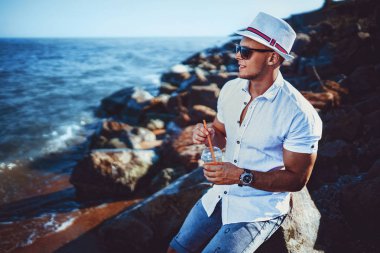 Tatil beldesinde beyaz gömlek ve şapka deniz arka planda bir kaya üzerinde oturan adam.