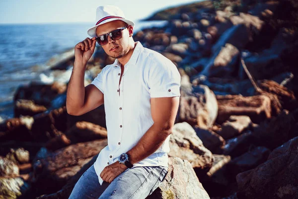 白いシャツと帽子をかぶったリゾートの男が海の背景の岩の上に座っている. — ストック写真