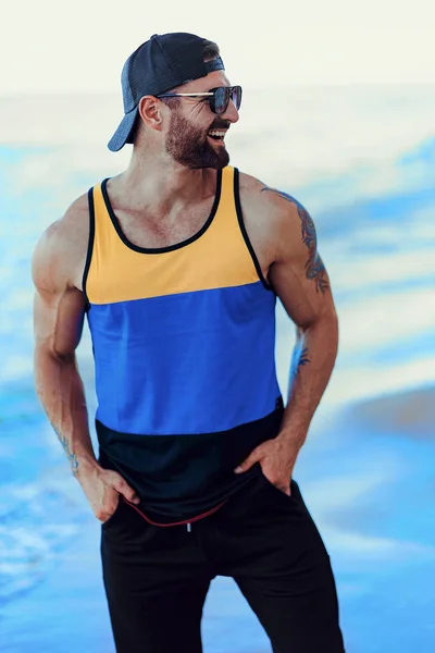 Brodaty tatuażem człowieka w ośrodku w Sportive shirt, czapka i okulary przeciwsłoneczne na tle morza — Zdjęcie stockowe