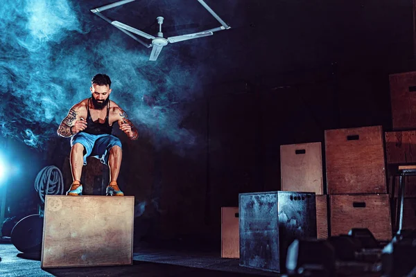 Fit tattoed bearded man jumping onto a box as part of exercise routine. Man doing box jump in the gym. Athlete is performing box jumps — Stock Photo, Image