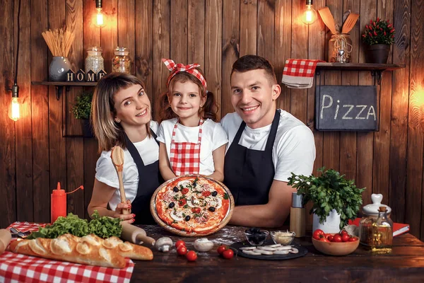 Familia feliz en delantales sonriendo y sosteniendo pizza cocinada —  Fotos de Stock