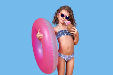 Mayo giymiş, güneş gözlüğü takan, elinde şişirilebilir yüzük ve mavi arka planda samanla hindistan cevizi tutan mutlu küçük bir kızın portresi.