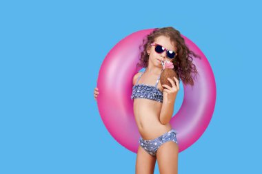 Mayo giymiş, güneş gözlüğü takan, elinde şişirilebilir yüzük ve mavi arka planda samanla hindistan cevizi tutan mutlu küçük bir kızın portresi.