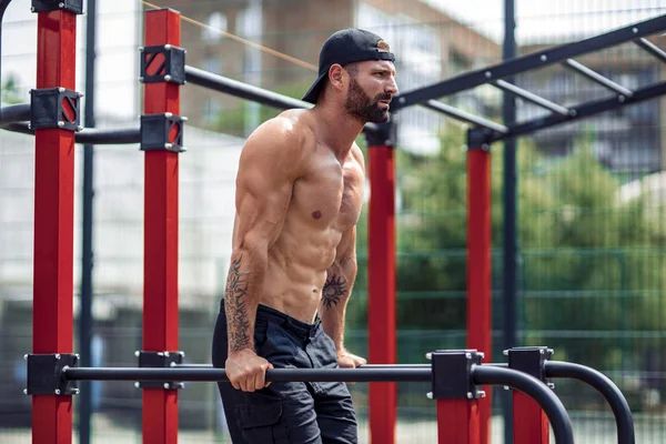 Сильный мускулистый мужчина отжимается в неровных барах в спортзале на улице. — стоковое фото
