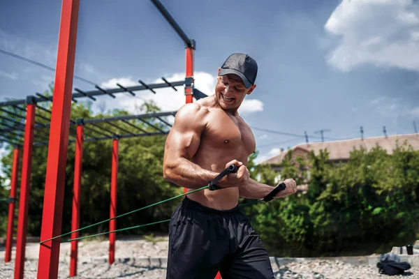 Homem de fitness treinando peito com bandas de resistência no quintal do ginásio de rua. Treino ao ar livre. Treino corporal com equipamento lá fora. Acessório elástico elástico elástico . — Fotografia de Stock