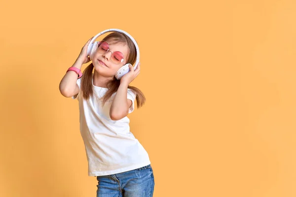 Meisje dat naar muziek luistert in een koptelefoon op een gele achtergrond. Leuk kind genieten van happy dance muziek, nauwe ogen en glimlach poseren op studio achtergrond muur — Stockfoto