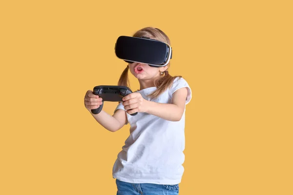 Menina experimentando VR headset vs joystick jogo no fundo amarelo. Emoções surpreendentes no rosto dela. Criança usando um gadget de jogo para realidade virtual . — Fotografia de Stock