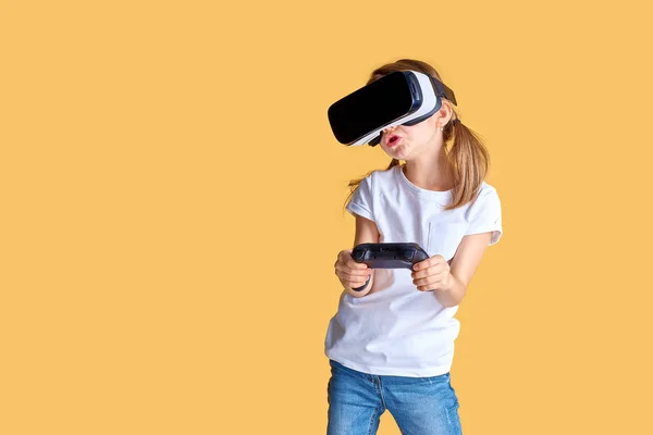 Menina experimentando VR headset vs joystick jogo no fundo amarelo. Emoções surpreendentes no rosto dela. Criança usando um gadget de jogo para realidade virtual . — Fotografia de Stock