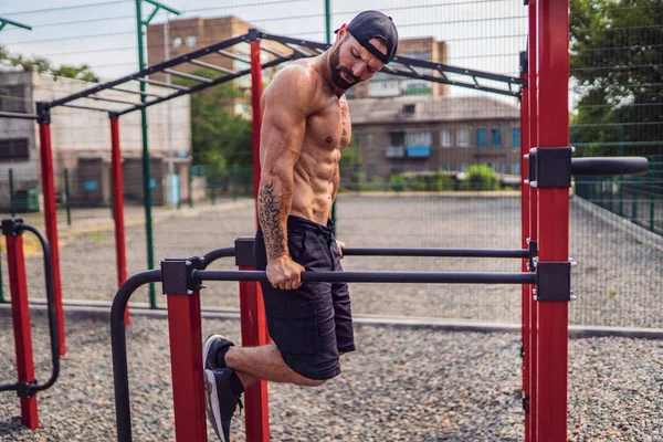 Сильный мускулистый мужчина отжимается в неровных барах в спортзале на улице. — стоковое фото