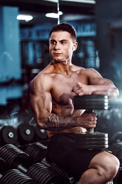 Young adult man handsome athlete working out in gym, sitting on a bench and holding dumbbell with raised arms. indoor, looking weight — Stock Photo, Image