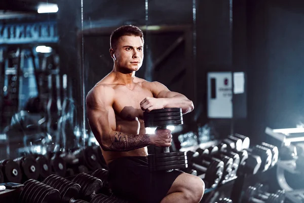 Νέος ενήλικος άντρας όμορφος αθλητής που εργάζονται έξω στο γυμναστήριο, καθισμένος σε ένα παγκάκι και κρατώντας αλά με ανυψισμένη χέρια. εσωτερικό, κοιτάζοντας το βάρος — Φωτογραφία Αρχείου
