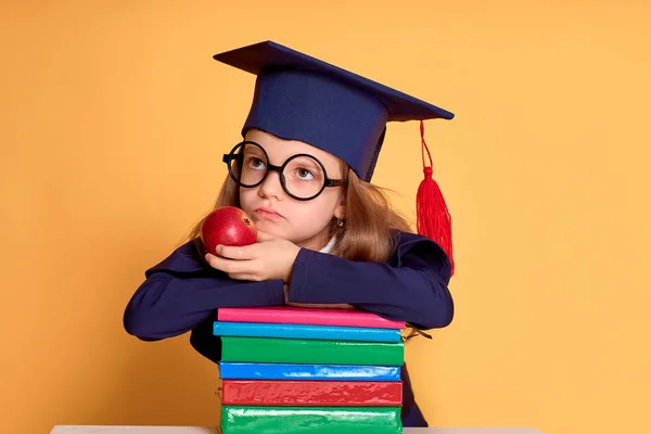 Menina pensativa em óculos e roupas de graduação pensando enquanto deitado sobre os livros coloridos — Fotografia de Stock