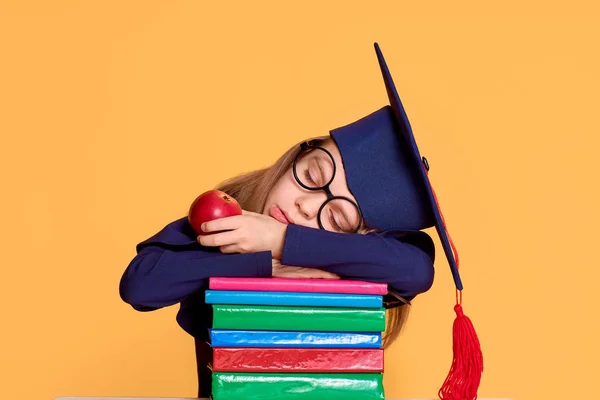 欢快的女学生在毕业服装下，拿着一个苹果在一堆课本上打趣 — 图库照片