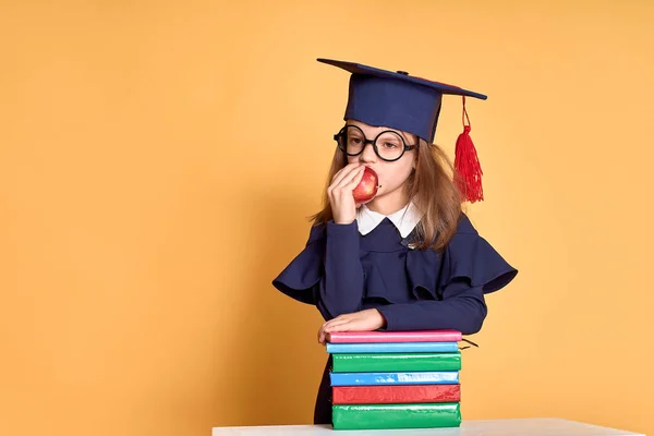 Vrolijke schoolmeisje in afstuderen outfit dragen Apple terwijl staan naast stapel van schoolboeken — Stockfoto