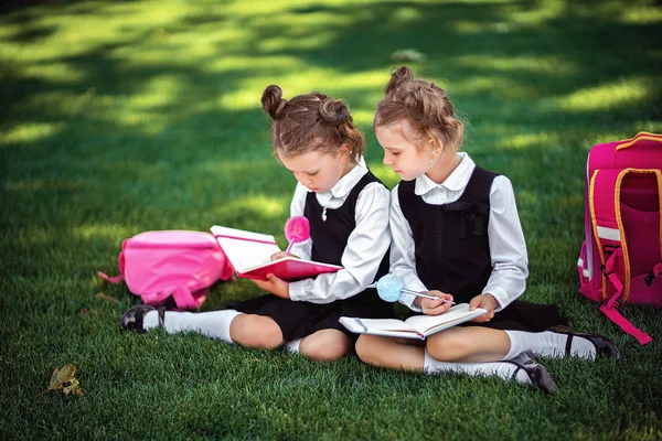 Two little school girls with pink backpack sitting on grass after lessons and read book or study lessons, thinking ideas, education and learning concept
