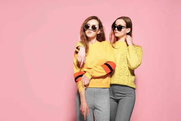 Two beautiful gorgeous girls standing in stylish yellow sweaters, gray pants, sunglasses on pink background. — Stockfoto