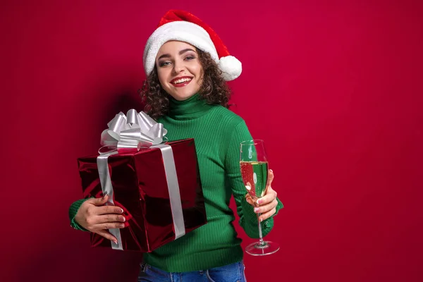 Beautiful fashion woman in santa claus hat, warm green Christmas sweater holding a glass of champagne and gift box on red background. — Stock Photo, Image