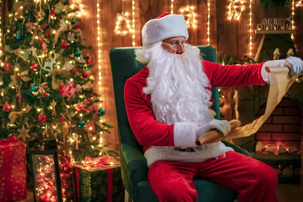 Porträt eines glücklichen Weihnachtsmannes, der zu Hause in der Nähe des Weihnachtsbaums sitzt und Weihnachtsbriefe oder Wunschzettel liest — Stockfoto