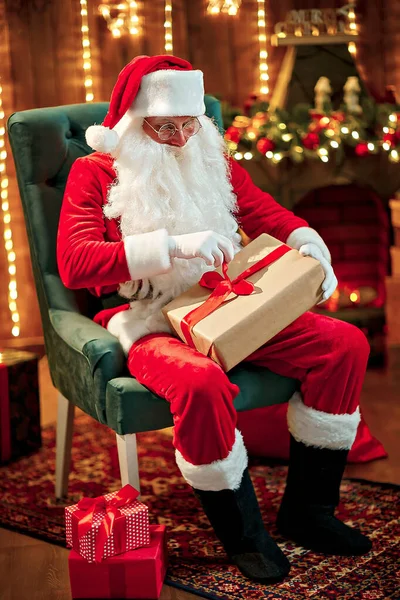 Foto de Papai Noel feliz em óculos sentados na cadeira, segure a caixa de presente — Fotografia de Stock