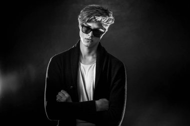 Siyah ceketli ve güneş gözlüklü havalı bir adam. Siyah arka planda poz veren yüksek moda erkek model..