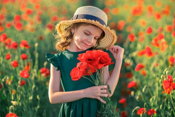 Yeşil elbiseli şirin küçük bir kız ve elinde bir buket haşhaş ile yaz günbatımında elinde haşhaş tarlasında poz veren hasır şapkalı. — Stok fotoğraf