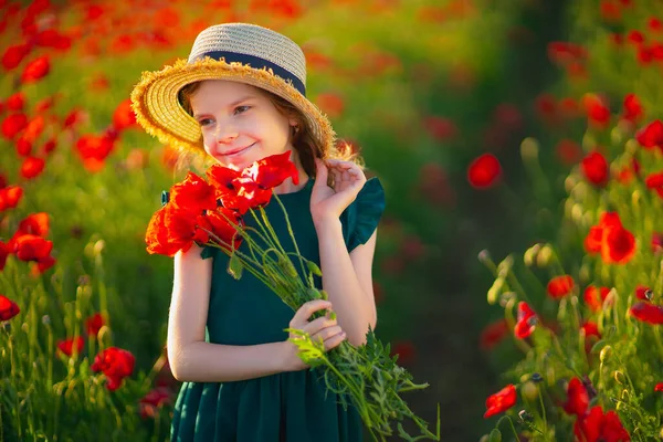 Cute little girl w zielonej sukience i słomy kapelusz z bukietem maków pozowanie na polu maków w jej rękach na letni zachód słońca — Zdjęcie stockowe