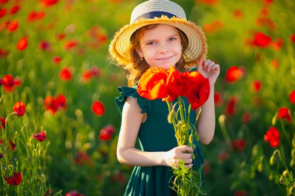 Schattig klein meisje in groene jurk en strohoed met een boeket klaprozen poseren op het veld van klaprozen in haar handen op de zomer zonsondergang — Stockfoto