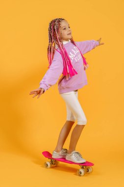 Aktif genç kız pembe gömlekli ve beyaz şortlu sarı arka planda kaykay tutuyor.