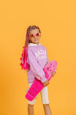 Aktif genç kız pembe gömlekli ve beyaz şortlu sarı arka planda kaykay tutuyor.