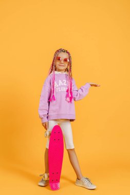 Aktif genç kız pembe gömlekli ve beyaz şortlu sarı arka planda kaykay tutuyor.