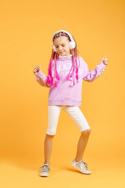 Yuvarlak gözlüklü sevimli bir çocuk pembe rasta takmış kulaklıkla müzik dinliyor ve sarı arka planda dans ediyor. Kulaklıkla müzik dinleyen sevimli küçük çocuk, kopyalama alanı.