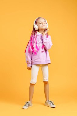 Yuvarlak gözlüklü sevimli bir çocuk pembe rasta takmış kulaklıkla müzik dinliyor ve sarı arka planda dans ediyor. Kulaklıkla müzik dinleyen sevimli küçük çocuk, kopyalama alanı.