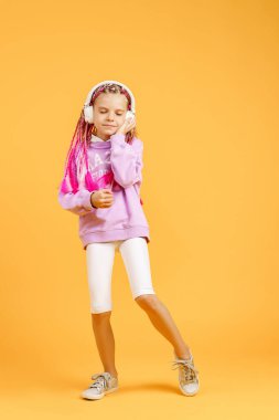 Yuvarlak gözlüklü sevimli bir çocuk pembe rasta takmış kulaklıkla müzik dinliyor ve sarı arka planda dans ediyor. Kulaklıkla müzik dinleyen sevimli küçük çocuk, kopyalama alanı.