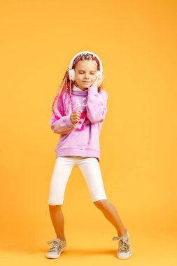 Yuvarlak gözlüklü sevimli bir çocuk pembe rasta takmış kulaklıkla müzik dinliyor ve sarı arka planda dans ediyor. Kulaklıkla müzik dinleyen sevimli küçük çocuk, kopyalama alanı.