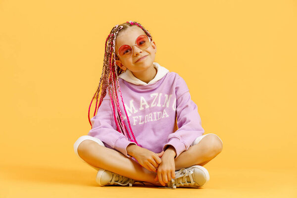 Stylish girl in rounded glasses with pink dreadlocks sitting, looking at camera and posing on a yellow background. Beauty, fashion