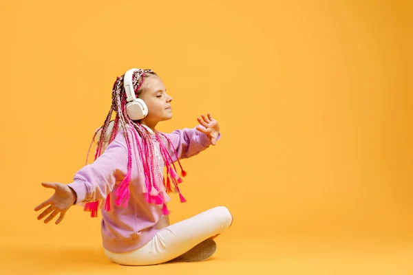 Vista posterior de Adorable niño en gafas redondeadas con rastas rosadas sentadas en el suelo y escuchar música tocando en auriculares sobre fondo amarillo. Lindo niño escuchando música en auriculares, espacio para copiar — Foto de Stock