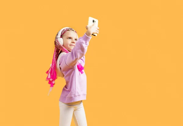 Pretty modern girl with pink dreadlocks in headphones smiling and making selfie on smartphone isolated over yellow — Stock Photo, Image