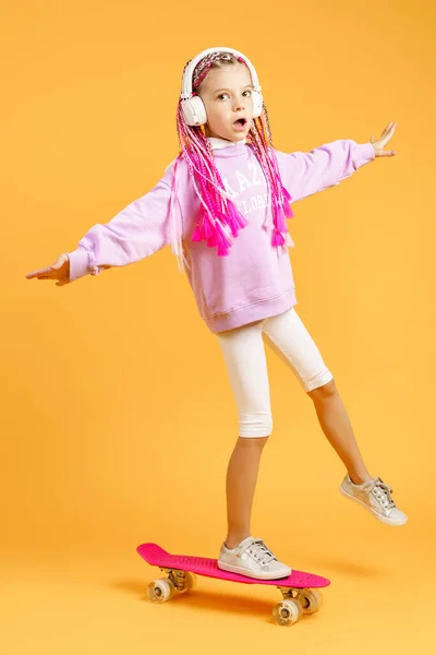 Menina ativa e feliz com cabelo encaracolado, fones de ouvido se divertindo com placa de centavo, smiling face stand skate. Penny board skate bonito para meninas. Menina passeio penny board amarelo fundo — Fotografia de Stock