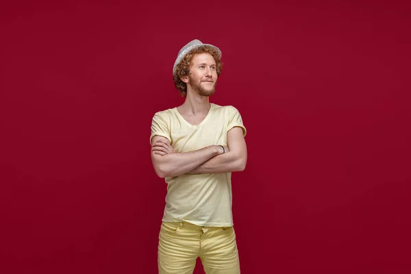 Hombre hipster de moda posando con los brazos cruzados en el estudio. Hombre sonriente en sombrero de paja mirando hacia otro lado de la cámara aislado en fondo rojo — Foto de Stock