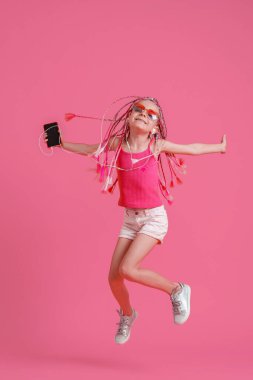 Rasta saçlı güzel bir kız elinde cep telefonuyla zıplıyor ve pembe arka planda kulaklıkla müzik dinliyor.