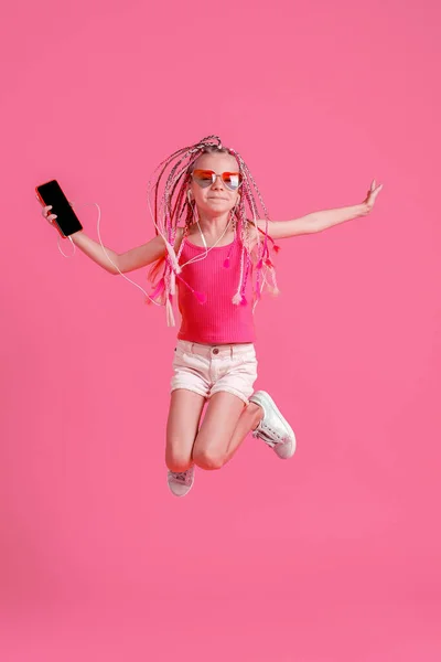 携帯電話でジャンプし、ピンクの背景にヘッドフォンで音楽を聴くドレッドロックと美しい女の子 — ストック写真