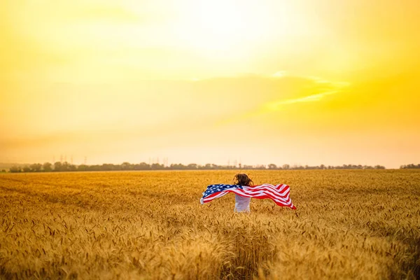 Mulher correndo e saltando despreocupado com os braços abertos sobre o campo de trigo Segurando bandeira dos EUA — Fotografia de Stock