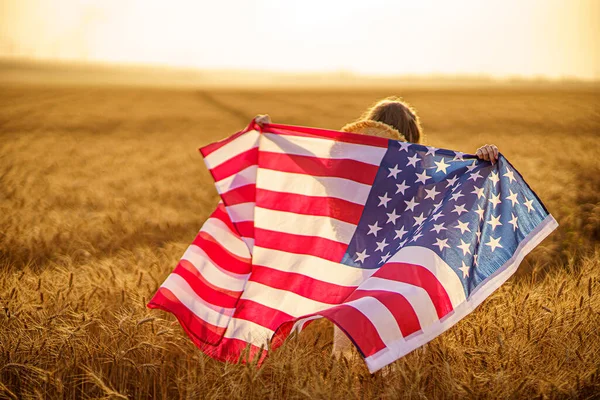 Vista trasera de una chica vestida de blanco con una bandera americana mientras corre en un hermoso campo de trigo — Foto de Stock