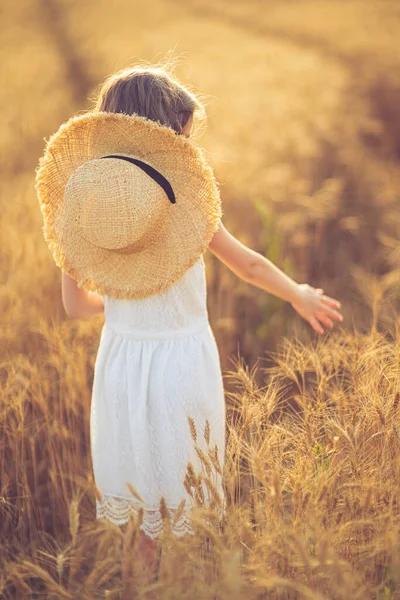 Buğday tarlasında beyaz elbiseli ve hasır şapkalı küçük bir kızın moda fotoğrafı. — Stok fotoğraf