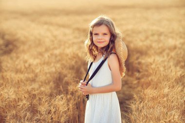 Buğday tarlasında beyaz elbiseli ve hasır şapkalı küçük bir kızın moda fotoğrafı.