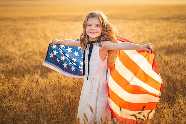 Adorável menina patriótica em vestido branco vestindo uma bandeira americana enquanto corre em um belo campo de trigo — Fotografia de Stock
