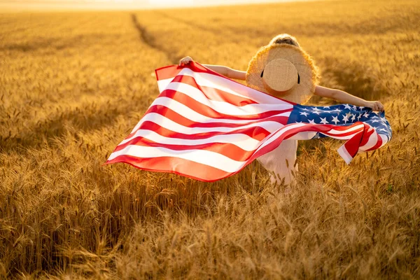 Visão traseira de uma menina de vestido branco vestindo uma bandeira americana enquanto corre em um belo campo de trigo — Fotografia de Stock