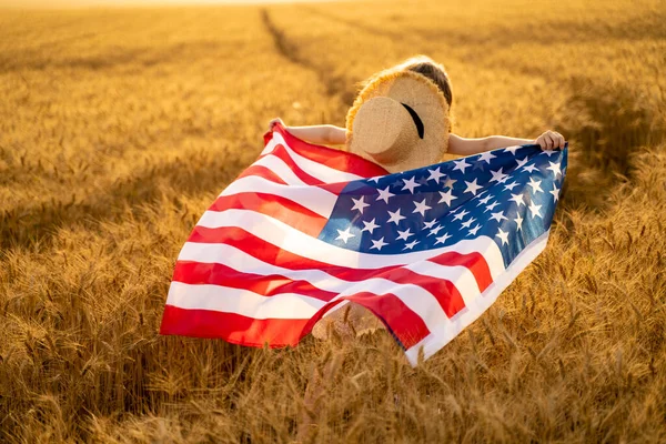 Visão traseira de uma menina de vestido branco vestindo uma bandeira americana enquanto corre em um belo campo de trigo — Fotografia de Stock